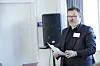 Mann står med et ark i handa og snakker, trolig til ei forsamling. Han har mørkt hår med gråstenk, briller, mørk skjorte og dressjakke.