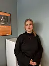 Ung kvinne i mørk genser står foran vegg med oransje plakat