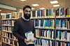 Student holder en stabel bøker foran bokhyller i bibliotek.