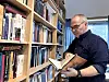 Mann leser i en bok. Foran seg har han en bokhylle med mange bøker.