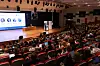 Fullsatt auditorium der publikum følger en presentasjon fra scenen. Bak innlederen er det en stor skjerm som viser hvem talerne er.