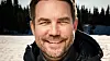 Smilende mann i vinterjakke foran solfylt snødekt landskap og skog.