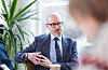 Mann i dress sitter på kontor med plante i bakgrunnen og en uskarp person i forgrunnen.