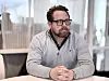 Mann med briller og skjegg sitter ved et bord i et lyst kontor.