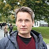 Mann i jakke står utendørs i en park med trær og mennesker i bakgrunnen.