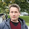 Mann i jakke står utendørs i en park med trær og mennesker i bakgrunnen.