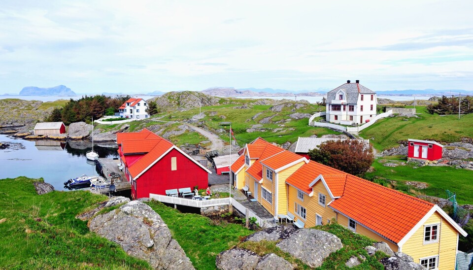 Bildet viser et landlig område. Et rødt, et gult og noen hvite hus spredt utover et stort gressområde. Flott natur. bak ser vi et gjell ute i havet.