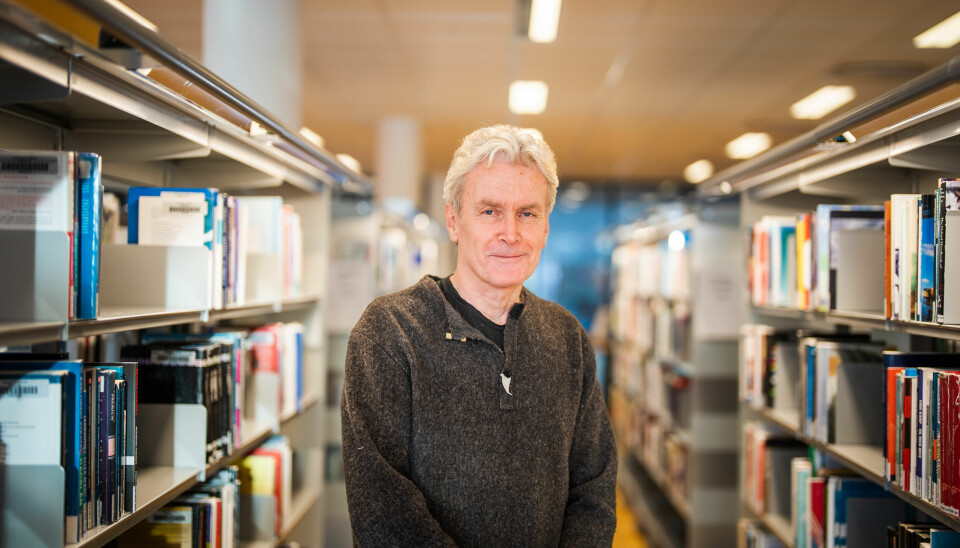 Bildet viser en mann med grått hår og et lite smil om munnen som står mellom noen bokyller i et bibliotek. Han ser rett inn i kameraet. Han har på seg en brun ullgenser og har hendene foldet.