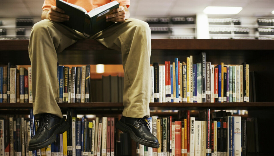 Mann leser bok mens han sitter på toppen av en bokhylle på et bibliotek.
