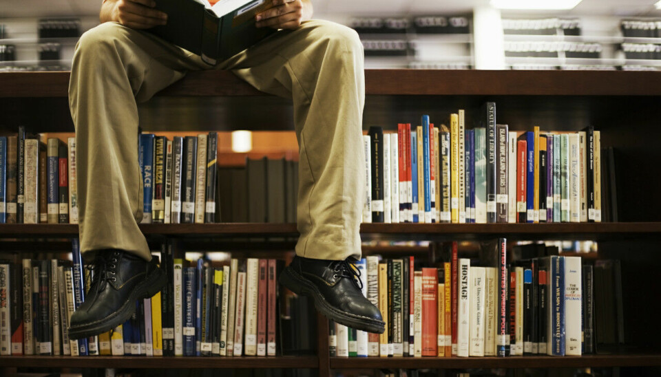 Mann leser bok mens han sitter på toppen av en bokhylle på et bibliotek.