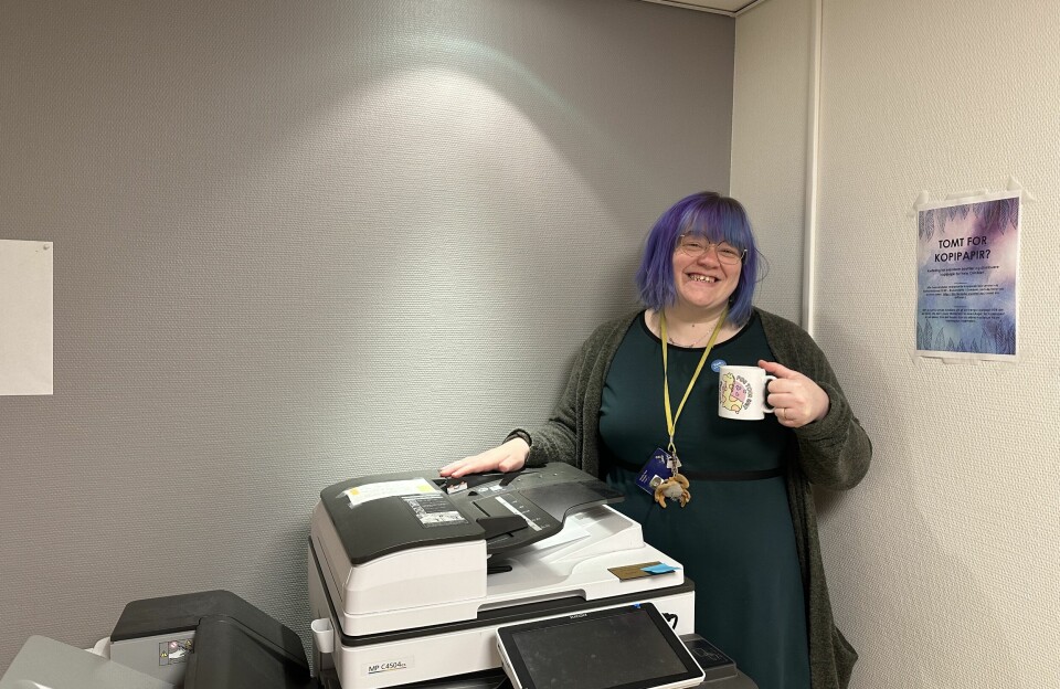 Camilla Soelseth, med grønn kjole og lilla hår, står med et smil og en kaffekopp ved siden av printeren.