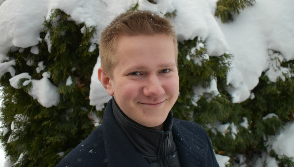 En smilende blond mann står foran noen furutrær dekket i snø.