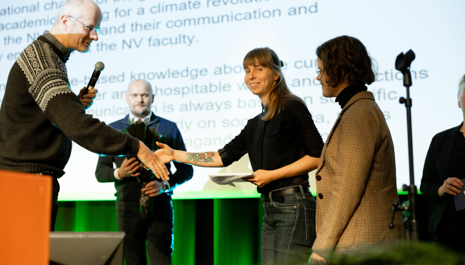 Øyvind Gregersen, i strikkegenser, holder ut hånda til Kristiina Visakorpi, en kvinne med svart genser og brunt hår som smiler mot kamera. De står på en scene med en skjerm full av skrift i bakgrunnen.