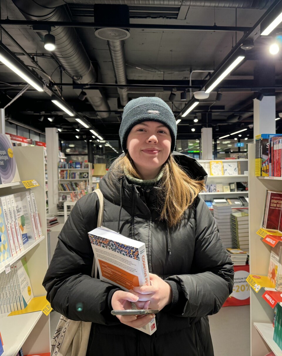 En ung kvinne med langt blondt hår, boblejakke og lue står mellom to bokhyller i en bokhandel. Hun har en bok i hendene og smiler.