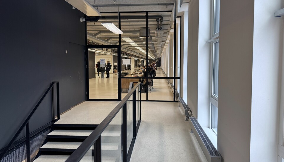 Et bilde med en trapp i forgrunnen og en korridor sett gjennom en glassdør og glassvegg. Det er mennesker som står i korridoren, og noen som sitter ved pulter.