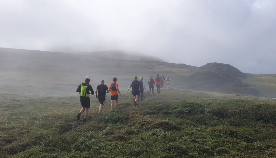 Et tåkelagt fjell med mange løpende sporty kledde mennesker med ryggen til kameraet mens de løper i maraton. Det er mose og grønt på bakken