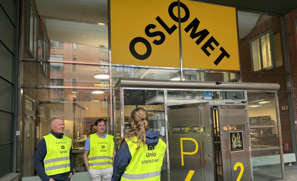 Tre menn står med refleksvest med teksten 'Unio Streikevakt' foran et bygg med logoen til 'OsloMet' med gul bakgrunn