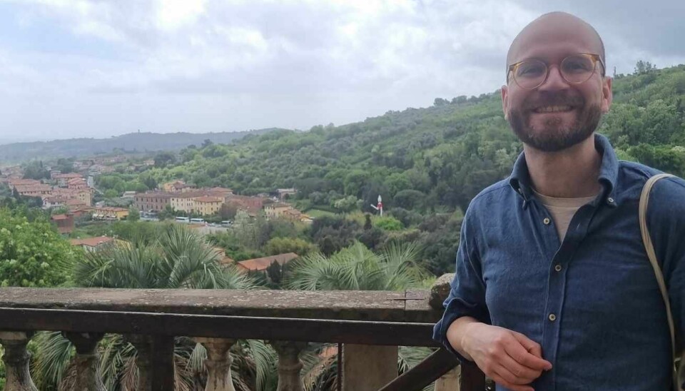 Smilende mann i blå skjorte, på en terrasse med utsikt over palmer og en landsby i fjellene